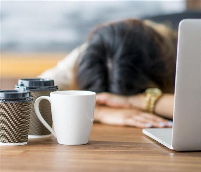 A lady slumped over her desk feeling fatigued. She has her laptop, with cups of coffee.